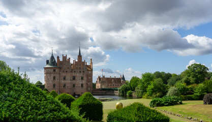 Egeskov Castle, a historic Renaissance water castle in Denmark, stands surrounded by its formal gardens, lush greenery, and a reflecting moat on a cloudy day © photoopus
