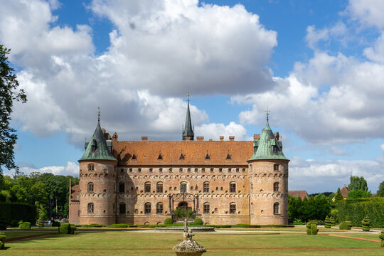 Egeskov Castle. A preserved Renaissance water castle. Featuring its red brick facade. Green copper turrets. And pointed spire under a blue sky with white clouds. Surrounded by well-maintained gardens