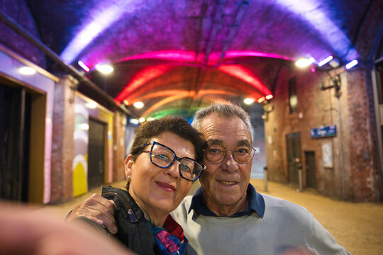 Older couple embracing and smiling for a selfie in a colorful, illuminated urban archway at night, capturing a joyful travel moment of togetherness and authentic connection