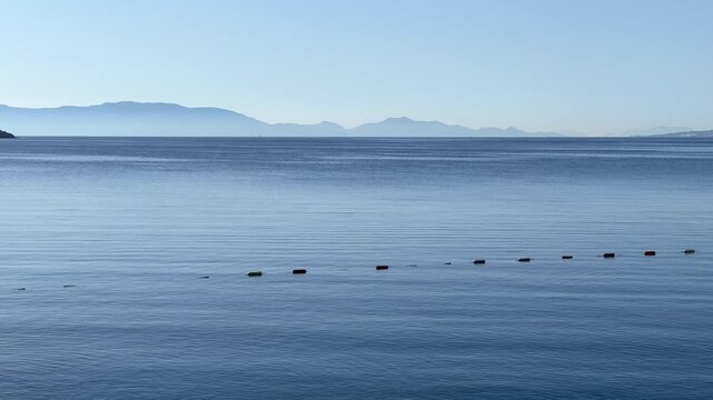 A tranquil panoramic view of calm blue sea water with a line of safety buoys and distant hazy mountains under a clear sky.