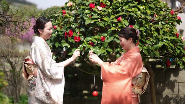 Two adult women in patterned kimono face each other outdoors while handling a red ball attached to a string beside a flowering shrub.