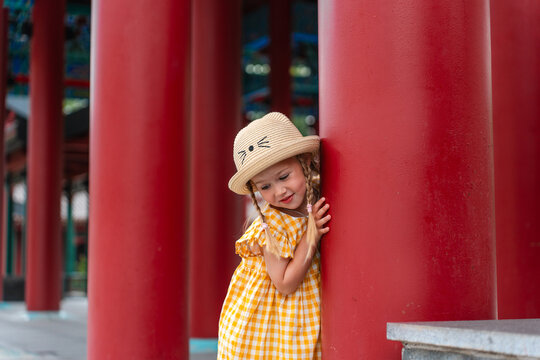 A close-up portrait of a cute little blonde girl with braided pigtails wearing a straw hat with cat ears and a yellow checkered dress with ruffled sleeves.