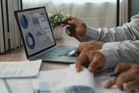 Two businessmen discussing financial data during business meeting, reviewing laptop charts and printed reports with pen for financial analysis and performance reporting.