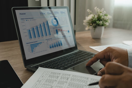 Businessman analyzing financial data on laptop dashboard while reviewing printed spreadsheet report with pen, showing accounting review, planning and business reporting.