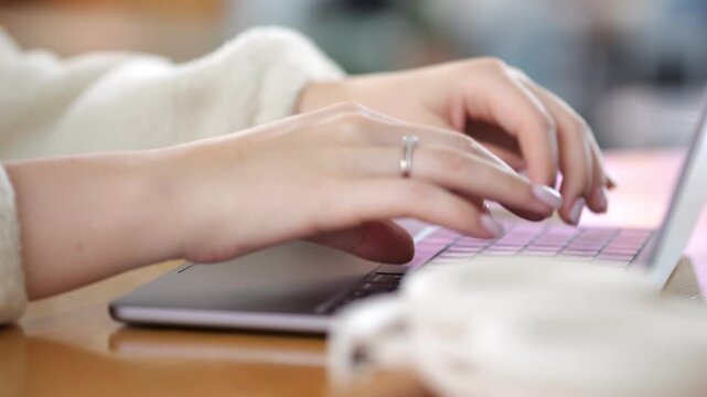 Close-up of hands typing on laptop keyboard. Fingers move quickly over keys. Ring visible on left hand. Soft focus background shows cozy indoor setting. Warm wooden table surface beneath device