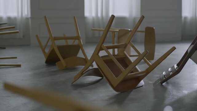 Overturned wooden chairs scattered across an empty grey panelled room in cold light