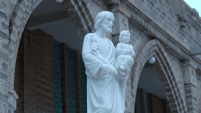 A white marble statue of Saint Joseph holding the infant Jesus, positioned near a stone architectural archway of the Christ the King Cathedral (also known as the Stone Church or Nha tho Nui) in Nha Tr