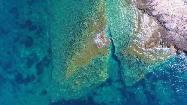 Aerial drone overhead of reef and shallow underwater rocks near a rocky peninsula and islet.