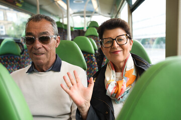 Senior couple enjoying their journey on public transport, a smiling woman waving at the camera with her husband sitting next to her on the green seats of the bus © Fotografia Juan Reig
