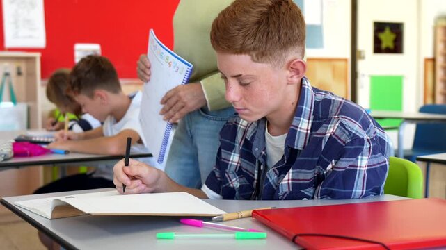 Attentive redhead boy with freckles sitting at his desk in a classroom, focused on writing in his notebook while the teacher supervises the students during a lesson at school
