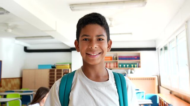 Cheerful elementary school student wearing a backpack standing inside a bright, colorful classroom and smiling confidently at the camera, ready for a new day of learning and education