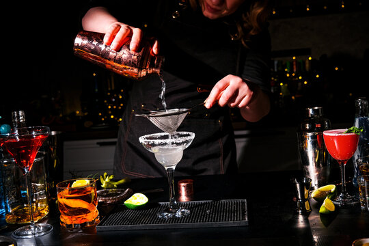 Female bartender pouring margarita cocktail drink through strainer into glass. Expert mixology and nightlife entertainment. Woman preparing cold tequila alcoholic beverage at dark bar.