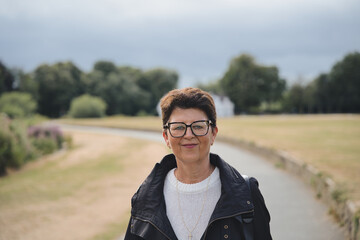 Mature woman with glasses and short brunette hair stands confidently on a paved park path in yorkshire, smiling in a casual white sweater and black jacket on a sunny day © Fotografia Juan Reig
