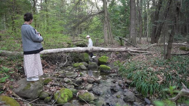 Two adult women are positioned near a shallow rocky stream in woodland, with one standing in the foreground and another farther back.