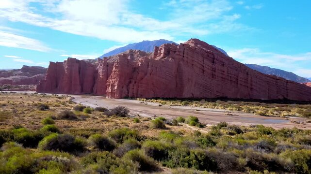 Los Castillos sandstone cliffs rise above a dry riverbed and desert shrubs in Quebrada de las Conchas during a daytime drone flyover.