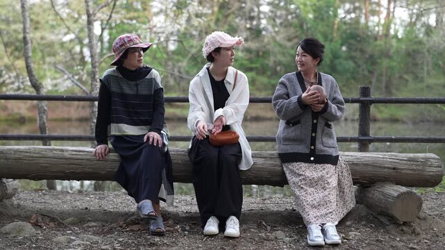 Three adult women sit side by side on a rustic log bench surrounded by trees.