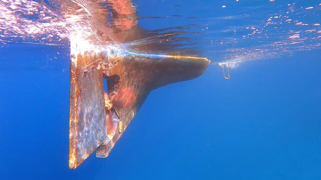 A boat propeller and rudder appear underwater beneath the stern in clear blue water.