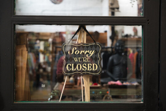 Closed sign with sorry we're closed message hanging on a dark framed shop window, reflecting the street and showing a blurry store interior with clothes and a buddha statue