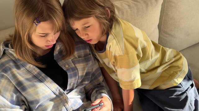 Two kids lean over together on a light sofa, attentively watching video content on a smartphone, modern home.