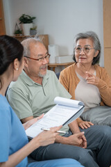 A home health nurse reviews a medical checklist on a clipboard with a senior Asian couple sitting...