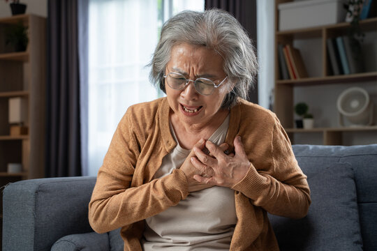 Senior Asian woman sitting on a sofa at home experiencing sudden chest pain, clutching her chest with both hands, eyes closed and face grimacing in discomfort.