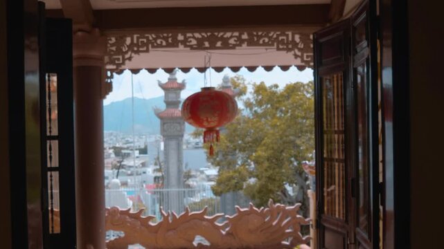 A big red Chinese silk lantern sway on the wind near intricately carved Vietnamese stone columns with tiled roofs against the background of the hills and houses of Nha Trang City in Vietnam. Shot in m