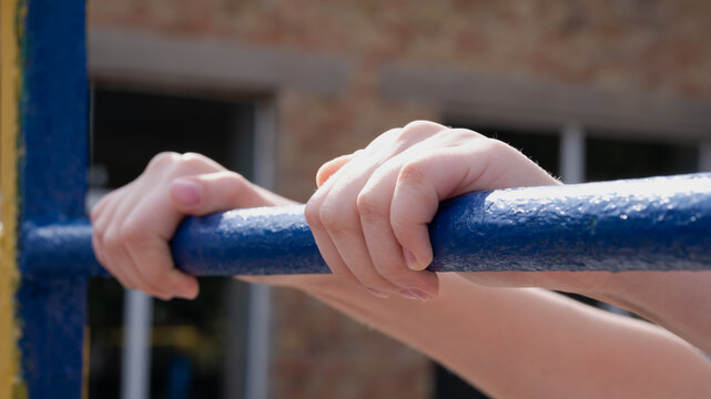 Close-up of a child's hands gripping a blue metal horizontal pull-up bar outdoors. Active kid practicing pull-ups on sports playground with blurred background and sunny daytime lighting.