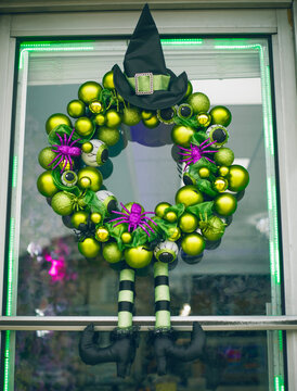 Halloween witch wreath decoration hanging on a glass door. Spooky green bauble wreath featuring a black witch hat, striped legs, purple spiders, and creepy eyeballs, festive autumn holiday home decor.