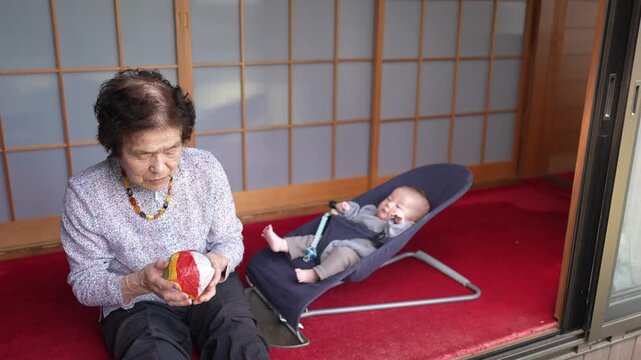 Older woman looks down at a colorful patchwork ball in her hands while a baby reclines beside her in a bouncer.