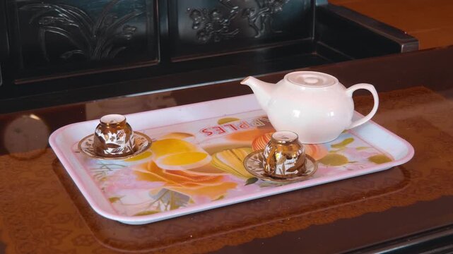 A set of a white ceramic tea pot and two upside down patterned teacups on saucers stands on a white lightweight printed melanin tray. Shot in motion
