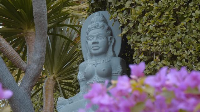 An ornate stone statue of an Asian goddess or deity, in Cham-style features a detailed headdress and calm face is placed outdoors among green plants and flowers. Shot in motion
