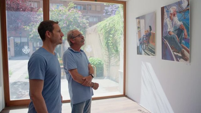 A senior man and a young man stand side by side near a bright window looking at a framed painting, Slow Motion.