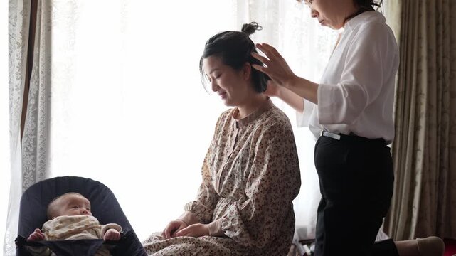 Seated adult woman has her hair arranged by another adult while a baby rests in a bouncer beside them near a bright window.