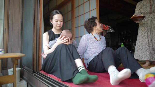 Younger woman in black clothing holds a baby while seated beside an older woman at an open doorway.