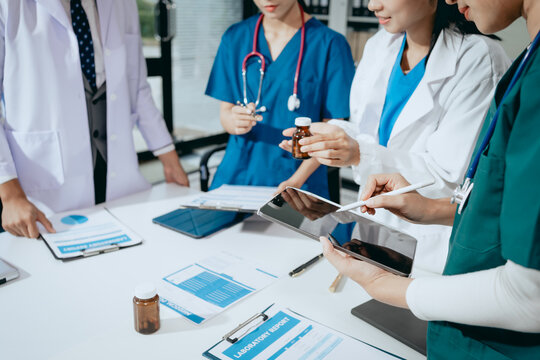 Medical team in white coats and scrubs reviews medicine bottles, tablets, charts and digital devices during a hospital meeting.