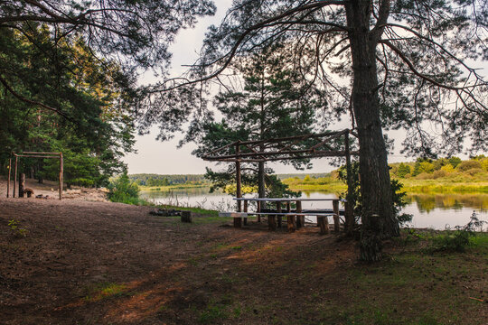 Rustic picnic area by the river in pine forest. Handmade wooden table and benches sheltered by rustic canopy under pine trees. Outdoor recreation, eco-tourism, camping.