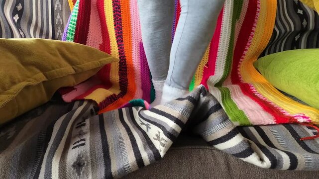 A person in comfy white socks and grey leggings leisurely moves their feet on a soft sofa, surrounded by colorful, striped blankets, embodying warmth and home comfort.