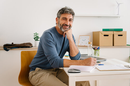 Friendly professional hispanic man writing documents at home desk. Portrait of mature freelancer looking at camera