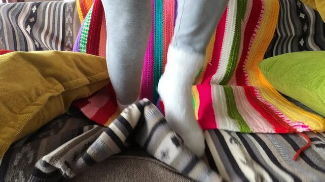 Cozy white fluffy socks move playfully, stepping on and off a stack of colorful striped socks, with a vibrant, handmade crocheted blanket providing a cheerful backdrop.