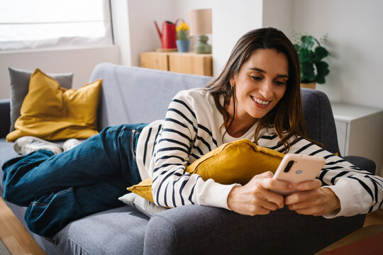 Smiling woman relaxing on a sofa at home using mobile phone app to share content on social media
