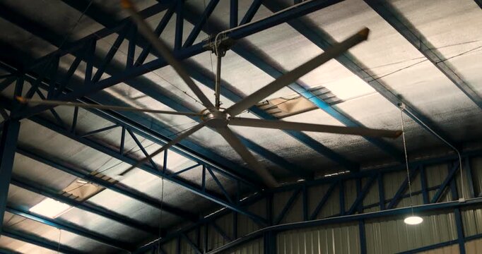 Large industrial ceiling fan spinning beneath a steel roof structure, ventilation and airflow, cooling, air circulation, warehouse and factory environment