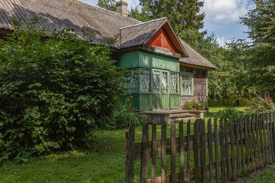 Сharming old wooden rural house with green porch surrounded by lush garden and wooden fence on summer day. Countryside life. Rural heritage, serenity, simplicity, beauty of traditional architecture.