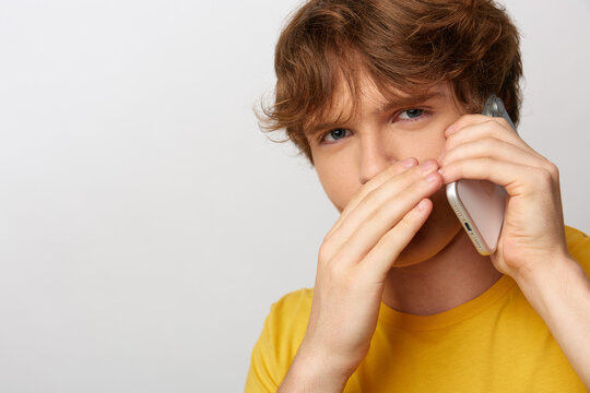 Suspicious young man in a yellow t-shirt covers his lower face while talking on a smartphone, listening to a questionable phone call with a wary, doubtful expression. Concept of scam and distrust.