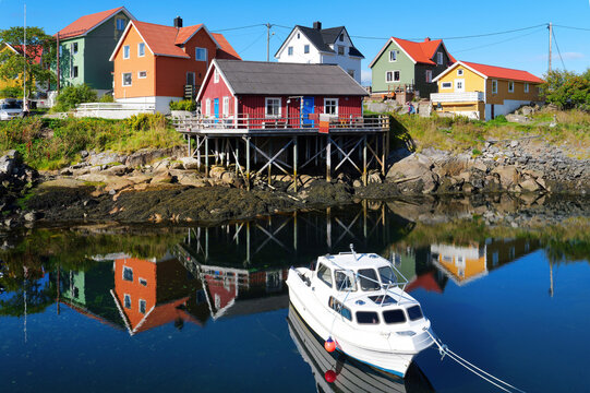 Colorful traditional wooden houses and a red building on stilts shape the coastal scene in Henningsv&aelig;r on the Lofoten Islands, while a white boat and houses reflect in calm water.
