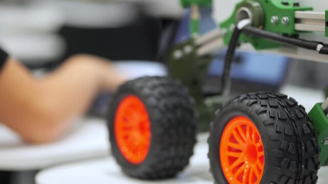 Close-up of orange-wheeled remote-controlled car. Person's hand adjusts green mechanical chassis. Background shows workshop with tools and computer