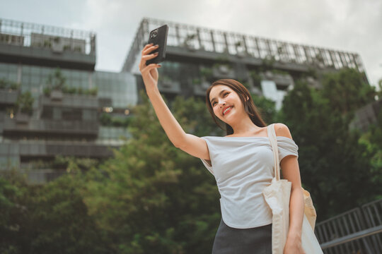Smiling young asian woman taking selfie with smartphone outdoors in modern city while carrying reusable tote bag, enjoying mobile technology, urban lifestyle, leisure and sustainable