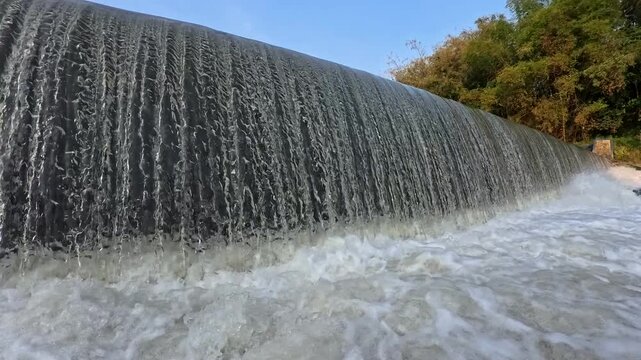 Close up of rushing water curtain overflowing from river weir dam into foamy riverbed