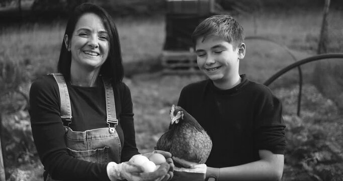 Happy mother and son holding chicken and eggs inside vegetable home garden - Family people having fun in fall time - Black and white editing - UGC content