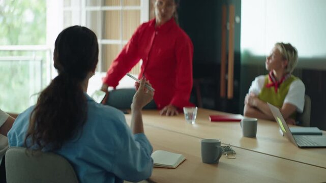 Coworkers gather around a conference table as a woman leads a business meeting in a bright office, presenting ideas and guiding team collaboration, discussion, and workplace communication.