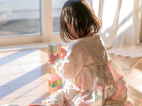 Little girl with short bob hair playing with pastel wooden blocks on the floor by a sunny window, back view cinematic shot
